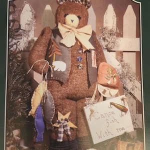 May include: A brown teddy bear wearing a vest and a bow tie, holding a fishing rod and a sign that says "Canoe fish with me". The bear is standing in front of a white picket fence and a green bush.