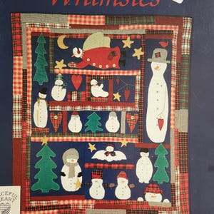 May include: A fabric wall hanging titled Winter Whimsies by Suellen Cochrane Wassem. The quilt features a dark blue background with appliqued snowmen, green pine trees, yellow stars, red hearts, and a flying angel, framed by a plaid patchwork border.