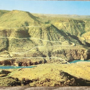 May include: A vintage postcard featuring a scenic view of a winding road through a mountainous landscape. The image showcases a river, green hills, and a clear blue sky. The road curves through the terrain, offering a picturesque view.