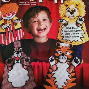 May include: A craft kit titled "Circus Puppets" featuring four colorful, stitched puppets: a clown, a lion, a dog, and a tiger. The puppets are made using 7-count plastic canvas. The kit includes a complimentary story.