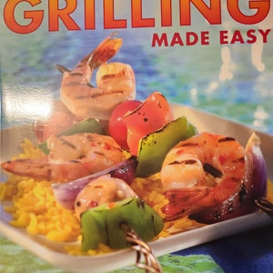 May include: A cookbook titled "Betty Crocker Grilling Made Easy" with a photo of a skewer of grilled shrimp, peppers, and onions on a bed of yellow rice. The book promises 200 sure-fire recipes from America's most trusted kitchens.