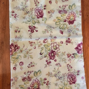 May include: Cream-colored fabric bag with a floral pattern featuring burgundy and pink flowers, green leaves, and small purple accents. The bag has a drawstring closure with a burgundy cord.