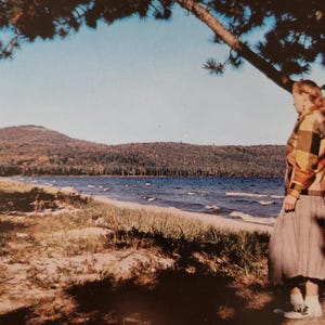 May include: A woman in a yellow and brown plaid jacket and a brown skirt stands on a sandy beach with a lake and forested hills in the background.