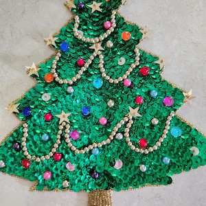 May include: A Christmas tree decoration made of green sequins, adorned with colorful beads and star-shaped embellishments. The tree has a gold beaded trunk and gold trim, creating a festive and sparkly appearance. The tree is a holiday decoration.