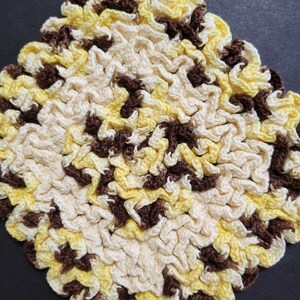 Puede incluir: Un posavasos de ganchillo con forma de diamante. El posavasos está hecho con hilo de color marrón, amarillo y crema.