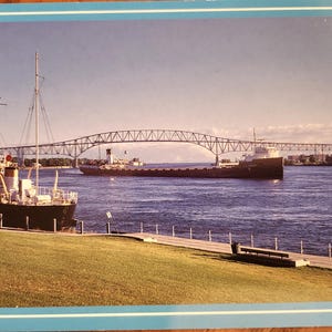 May include: A scenic photograph of a large bridge spanning a river, with a ship passing underneath. A smaller boat is docked on the left, and a grassy area and bench are in the foreground. The sky is blue.
