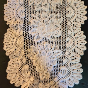 May include: A white lace table runner featuring an intricate floral pattern throughout the center and a scalloped edge design. The fabric has a delicate, open-weave texture with detailed needlework forming rose motifs and geometric borders.