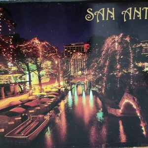 Puede incluir: Una postal que muestra una vista nocturna del San Antonio River Walk, iluminado por hileras de luces cálidas sobre árboles y edificios. Un barco flota en el agua oscura, reflejando las luces, con el texto SAN ANTONIO en la parte superior.