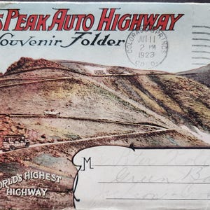 May include: Vintage postcard titled "Pikes Peak Auto Highway Souvenir Folder." The image depicts a winding road ascending a mountain, with vehicles visible. The text "The World's Highest Highway" is at the bottom. A postage stamp and a postmark from July 11, 1923, are also present.