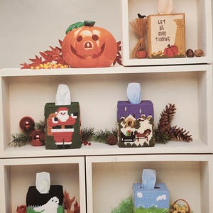 May include: Six different tissue box covers with various holiday themes. The covers are made of plastic canvas and feature cross-stitch designs. The designs include a pumpkin, a Thanksgiving scene, Santa Claus, a winter scene, a ghost, and a bunny rabbit.