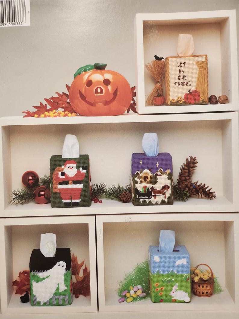 May include: Six different tissue box covers with various holiday themes. The covers are made of plastic canvas and feature cross-stitch designs. The designs include a pumpkin, a Thanksgiving scene, Santa Claus, a winter scene, a ghost, and a bunny rabbit.