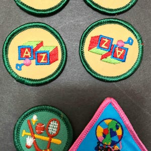 May include: Five green and yellow embroidered Girl Scout badges with the letters A and Z on blocks, and one pink and blue embroidered badge with the text "STEP FORWARD COOKIES 2000" and a globe with people running around it.