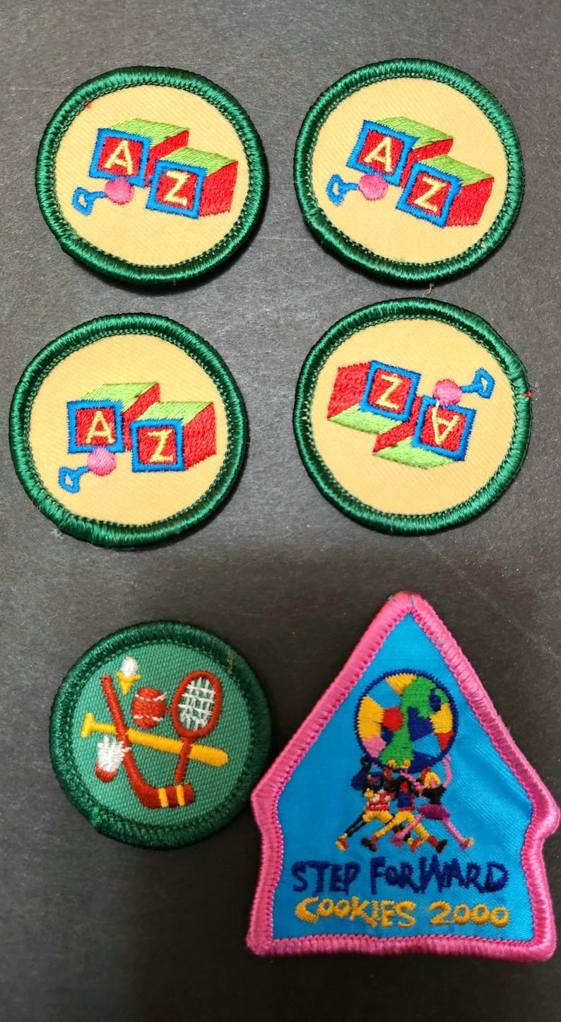 May include: Five green and yellow embroidered Girl Scout badges with the letters A and Z on blocks, and one pink and blue embroidered badge with the text "STEP FORWARD COOKIES 2000" and a globe with people running around it.