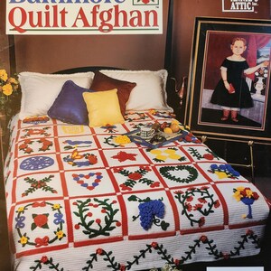 May include: A crochet pattern book titled "Baltimore Quilt Afghan" featuring a colorful, handmade quilt on a bed. The quilt has a white background with red borders and various floral and geometric designs. A framed portrait hangs on the wall.