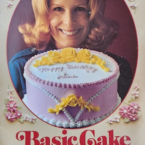 May include: A softcover instructional book titled Basic Cake Decorating The Wilton Way. The cover features a circular inset showing a person smiling behind a round cake with pink frosting, yellow icing roses, and the text Happy Birthday Sue.