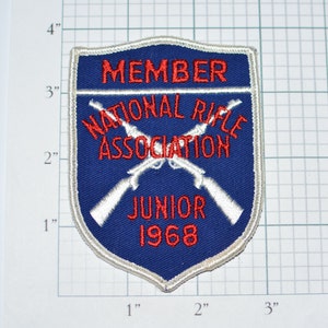 May include: A blue and white embroidered patch with the text "MEMBER NATIONAL RIFLE ASSOCIATION JUNIOR 1968". The patch features two crossed rifles in white.