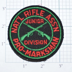 May include: A black, green, and red embroidered patch with the text "NAT'L RIFLE ASS'N JUNIOR DIVISION PRO-MARKSMAN" and a pair of crossed rifles.