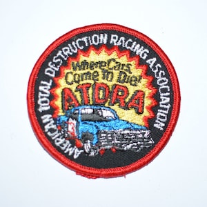 May include: A red, white, and blue embroidered patch with the text "American Total Destruction Racing Association" and "Where Cars Come To Die! ATDRA". A cartoon car is in the center of the patch.