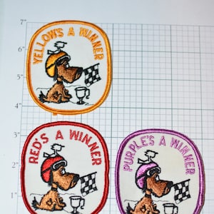May include: Three embroidered iron-on patches featuring a cartoon dog wearing a racing helmet and holding a checkered flag. The patches are oval shaped and have a white background with a colored border. The text on the patches reads "Yellow's a Winner", "Red's a Winner", and "Purple's a Winner".