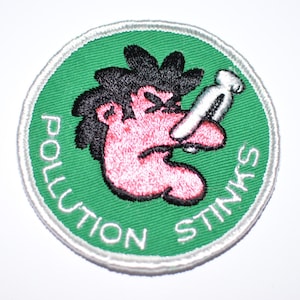 May include: A green and white circular patch with a cartoon illustration of a person with a red face and black hair holding their nose. The text "Pollution Stinks" is written around the edge of the patch.