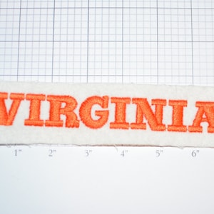 May include: A rectangular white patch with the embroidered word "VIRGINIA" in orange block letters. The patch measures approximately 7 inches long and 1 inch high.