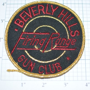 May include: A circular, gold-trimmed patch with black fabric and red embroidered text. The text reads "Beverly Hills Firing Range Gun Club".