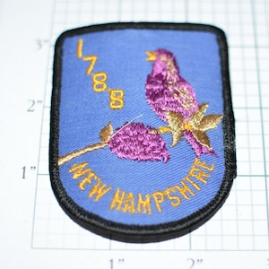 May include: A blue embroidered patch with a purple bird perched on a branch with purple flowers. The patch has the text "1788 NEW HAMPSHIRE" in yellow thread.