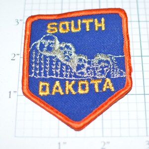 May include: A blue, shield-shaped patch with an orange border. The patch features the words "SOUTH DAKOTA" in gold lettering, with an embroidered image of Mount Rushmore in the center. The patch measures approximately 3 inches by 2 inches.