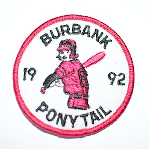 May include: A white embroidered patch with a red border. The patch features a girl in a red baseball uniform holding a bat. The text "Burbank Ponytail 19 92" is embroidered in black.