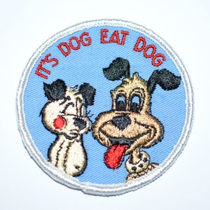 May include: A blue and white embroidered patch with two cartoon dogs. The text "It's Dog Eat Dog" is written in red around the edge of the patch.