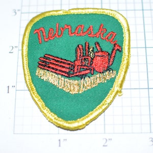 May include: A green and gold embroidered patch with the word "Nebraska" in red lettering. The patch features a red tractor with a gold plow, and the tractor is surrounded by a gold border.