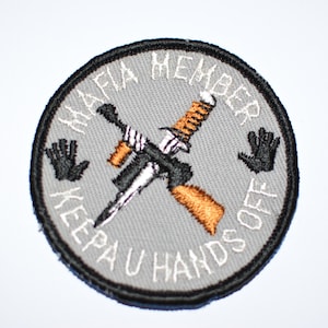 May include: A gray circular patch with black stitching around the edge. The patch has white text that reads "Mafia Member Keep Your Hands Off" with a crossed knife and gun in the center. The knife is silver and the gun is brown.