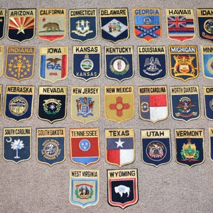May include: A collection of 50 embroidered patches, each representing a different state in the United States. The patches are arranged in rows, with the state name printed above each patch. The patches are all in the shape of a shield and feature various symbols and designs representing each state.