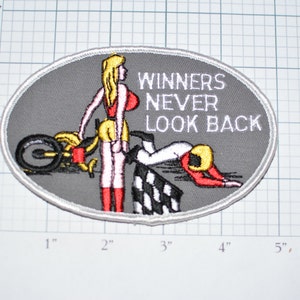 May include: A gray oval-shaped patch with a white border. The patch features a cartoon image of a woman in a red and yellow outfit standing next to a motorcycle. A man in a red and white racing suit is lying on the ground behind the woman. The text "WINNERS NEVER LOOK BACK" is printed in white on the patch.