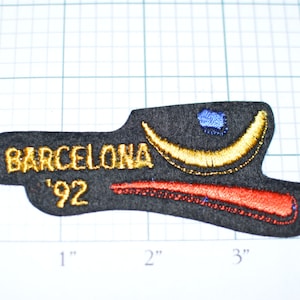 May include: Black felt patch with gold embroidered text "BARCELONA '92". The patch also features a gold crescent moon, a red and orange stripe, and a blue circle.