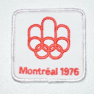 May include: A white embroidered patch with a red Olympic rings logo and the text "Montréal 1976".