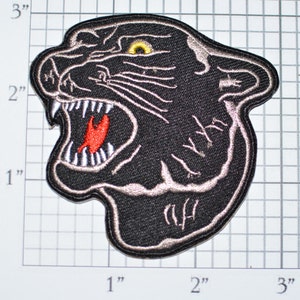 May include: Black embroidered iron-on patch of a snarling panther head with a yellow eye. The panther's mouth is open, showing sharp teeth and a red tongue.