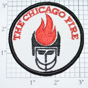 May include: A white, round patch with black stitching around the edge. The patch features a red flame with a black helmet in front of it. The text "THE CHICAGO FIRE" is written in a black circle around the flame and helmet.