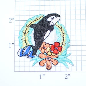 May include: An embroidered patch featuring a black and white orca whale jumping through a ring of tropical leaves and flowers. The flowers are red, orange, and yellow.