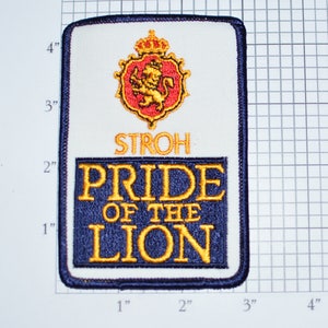 May include: A white embroidered patch with a blue border. The patch features a gold lion with a crown on a red shield. The text "STROH PRIDE OF THE LION" is in gold lettering on a blue background.