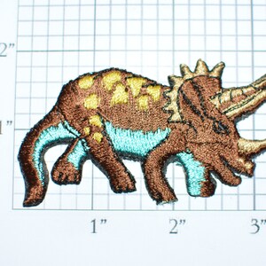 May include: Brown embroidered iron-on patch of a triceratops dinosaur with yellow and teal accents. The dinosaur is walking with its head turned to the side. The patch is approximately 2 inches wide.