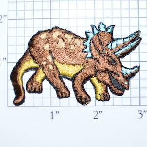 May include: Brown embroidered iron-on patch of a triceratops dinosaur with blue accents. The dinosaur is facing right and has three horns on its head.