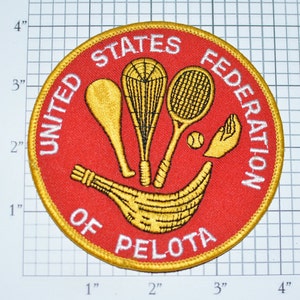 May include: A red and gold embroidered patch with the text "United States Federation of Pelota". The patch features a variety of sports equipment, including a tennis racket, a racquetball racket, a lacrosse stick, and a ball.