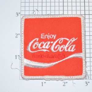 May include: A red fabric patch with white embroidered text that reads "Enjoy Coca-Cola Trademark" with a white wave design underneath.