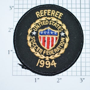 May include: A black circular patch with gold embroidery. The patch reads "Referee United States Soccer Federation 1994". The center of the patch features a shield with the American flag.