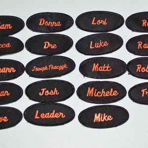 May include: Twenty black oval-shaped name tags with orange embroidered lettering. The names include Adam, Donna, Lori, Rae, Becca, Dre, Luke, Ramiro, Breann, Joseph Tkaczyk, Matt, Robert, Brian, Josh, Michele, Trent, Dave, Leader, and Mike.