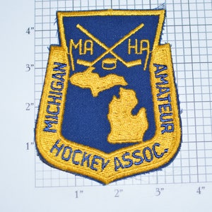 May include: A blue and gold embroidered patch with the text "Michigan Amateur Hockey Assoc." and a crossed hockey stick design. The patch also features an outline of the state of Michigan.