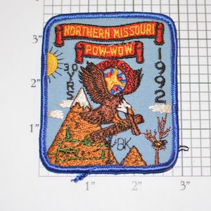 May include: A blue embroidered patch with a red banner that reads "Northern Missouri Pow-Wow 1992". The patch features a brown eagle with a yellow star on its chest, flying over a brown mountain with a white peak. The eagle is holding a red, white, and blue banner in its talons.