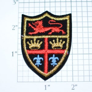 May include: A black embroidered patch with a gold border, featuring a red lion, a red cross, gold crowns, and blue fleurs-de-lis.