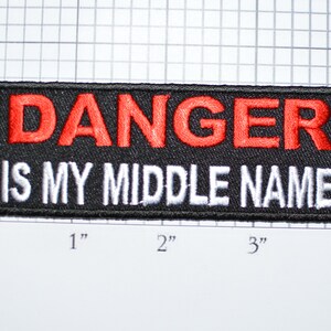 May include: Black embroidered patch with white and red lettering. The patch reads "DANGER IS MY MIDDLE NAME".